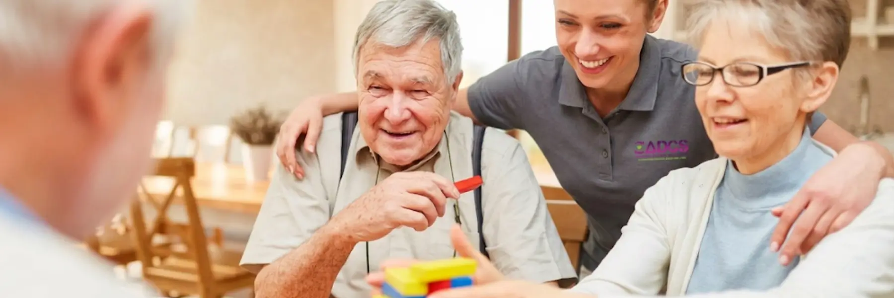 A caregiver smiles and embraces an elderly couple playing with colorful blocks at a table, creating a joyful and supportive atmosphere