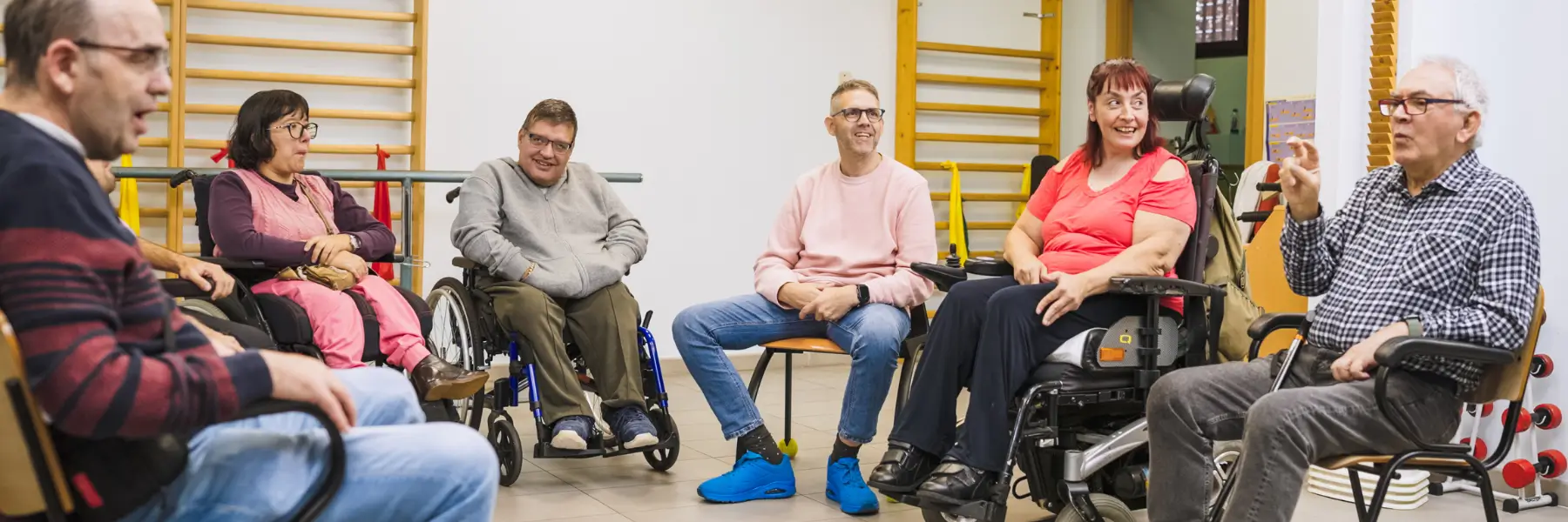 A diverse group of six people, some in wheelchairs, are seated in a circle in a bright room